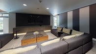 This single-floor media and lounge space is designed with a strong emphasis on comfort and cinematic experience. The room centers around a large wall-mounted flat-screen television flanked by warm wood-toned cabinetry with under-cabinet LED accent lighting, creating an inviting, theater-like ambiance. A generously sized sectional sofa in charcoal grey with cream accent pillows wraps around a substantial wood and concrete coffee table, providing ample seating for gatherings or screenings. One wall features a dramatic design element of bold vertical stripes in gold, taupe, and dark charcoal, adding visual depth and a contemporary aesthetic to the space. Recessed ceiling lighting supplements the ambient glow throughout the room. Neutral, textured carpet anchors the lounge area, complementing the overall dark and sophisticated color palette. A floating ledge shelf along one wall offers additional surface space, while a frosted-glass interior door with a warm wood frame provides a refined transition point. A window along one side introduces natural light, offering a subtle connection to the exterior. The overall design balances modern minimalism with warm, layered textures to create a polished and functional entertainment environment.