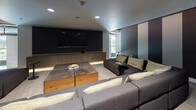 This single-floor media and lounge space is designed with a strong emphasis on comfort and cinematic experience. The room centers around a large wall-mounted flat-screen television flanked by warm wood-toned cabinetry with under-cabinet LED accent lighting, creating an inviting, theater-like ambiance. A generously sized sectional sofa in charcoal grey with cream accent pillows wraps around a substantial wood and concrete coffee table, providing ample seating for gatherings or screenings. One wall features a dramatic design element of bold vertical stripes in gold, taupe, and dark charcoal, adding visual depth and a contemporary aesthetic to the space. Recessed ceiling lighting supplements the ambient glow throughout the room. Neutral, textured carpet anchors the lounge area, complementing the overall dark and sophisticated color palette. A floating ledge shelf along one wall offers additional surface space, while a frosted-glass interior door with a warm wood frame provides a refined transition point. A window along one side introduces natural light, offering a subtle connection to the exterior. The overall design balances modern minimalism with warm, layered textures to create a polished and functional entertainment environment.