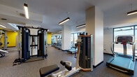 Located at 355 N Rosalind Ave in Orlando, Florida, this well-equipped fitness center offers a comprehensive workout environment across a single open floor. The space is designed with a modern aesthetic, featuring bold yellow accent walls paired with clean white surfaces, rubber flooring, and suspended linear lighting that ensures bright, even illumination throughout.

The strength training area includes a Smith machine, adjustable benches, a full rack of dumbbells displayed along a mirrored wall, and multiple selectorized machines for targeted muscle training. A dedicated functional training zone houses a wall-mounted rig with suspension trainers, resistance bands, and an organized rack of medicine balls and kettlebells, alongside spin bikes for cycling workouts. The cardio section is lined with treadmills and elliptical machines positioned near large windows, providing natural light and outdoor views of the surrounding Orlando skyline.

Additional highlights include a water fountain station conveniently located near the exit, wall-mounted televisions for entertainment during workouts, and ample open floor space that accommodates a variety of training styles. Structural columns are thoughtfully integrated into the layout without disrupting the flow of the space. The overall ambiance is energetic and functional, making this fitness center a well-rounded facility suited for a diverse range of fitness needs.