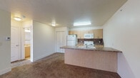 This single-floor residence is organized around a practical, open-concept layout that connects the kitchen and living areas seamlessly. The kitchen is equipped with light wood cabinetry, a dark stone-patterned backsplash, and white appliances including a refrigerator, electric range, and built-in microwave. A peninsula counter with a built-in sink and laminate facing extends into the living area, defining the boundary between the two spaces. The kitchen features light tile flooring, while the adjoining living area transitions to wall-to-wall carpeting that continues throughout the unit.

The living space is generously sized, with textured ceilings, neutral-toned walls, and access to a window that allows natural light. A separate bedroom offers carpeted flooring, a ceiling fan with light fixture, and a window with blinds providing natural illumination. A closet with bi-fold doors provides dedicated storage.

The bathroom is finished with a marble-look tile surround encasing the full tub-shower combination, a wood-framed vanity topped with stone-look counters, and tile flooring throughout. A dedicated laundry area features built-in shelving, washer/dryer connections, and a floor drain, offering convenient in-unit functionality.
