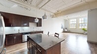 Located in Denver's vibrant RiNo neighborhood, this single-floor loft-style unit at 2101 Market St blends industrial character with modern finishes. The open-concept living and kitchen area is anchored by soaring ceilings with exposed white-painted wood beams, ductwork, and plank ceilings that lend a distinctly urban, warehouse-inspired aesthetic. Light hardwood flooring runs throughout the main living areas, reflecting natural light that pours in through large, yellow-trimmed windows.

The kitchen is a standout feature, equipped with dark espresso cabinetry, granite countertops, a classic subway tile backsplash, stainless steel appliances including a full-size refrigerator and dishwasher, and a generous island with bar seating — all illuminated by stylish pendant lighting. The spacious living area offers an open canvas with exposed structural elements overhead.

The bedroom is a bright, carpeted retreat with large windows framing city views, exposed ceiling beams, and a ceiling fan. It includes a walk-in closet outfitted with wire shelving on multiple walls for ample storage. Additional spaces include a carpeted secondary room with an open loft-like feel, a hallway with wood-look flooring, and well-appointed utility areas, completing this thoughtfully designed urban residence.
