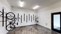 Located at 2131 N. Meridian, this dedicated bike storage room is a practical and well-organized amenity space designed to accommodate cyclists with ease. The room features a row of wall-mounted vertical bike racks lining the white-painted walls, allowing multiple bicycles to be stored securely in an upright position while maximizing floor space efficiency. The flooring consists of polished, sealed concrete with a distinctive marbled finish, offering a durable and easy-to-maintain surface well-suited for a high-use storage environment. Overhead, bright fluorescent strip lighting ensures the room is well-illuminated, making it easy to access and retrieve bicycles at any time of day. The clean white walls and minimalist design keep the space feeling open and functional. A small utility panel is visible along the lower wall, adding to the room's practical character. Overall, this bike storage space reflects a thoughtful approach to amenity design, prioritizing organization, durability, and convenience for residents or occupants who rely on cycling as part of their daily routine.
