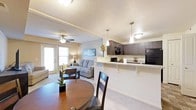 This single-floor residence offers a thoughtfully designed open-concept layout that seamlessly connects the kitchen, dining, and living areas into one cohesive living space. The kitchen is outfitted with dark cabinetry, black appliances including a refrigerator and microwave, and a light-toned countertop that extends into a breakfast bar with pendant lighting overhead — ideal for casual dining or entertaining. Adjacent to the kitchen, the dining area features a warm-toned round wooden table complemented by dark wood chairs, adding a touch of classic elegance. The living room is generously proportioned, anchored by plush seating and a ceiling fan that enhances comfort. Large windows and a glass door flood the space with natural light and provide access to an outdoor area, creating an airy, open feel throughout. The flooring transitions from rich hardwood in the dining zone to soft carpeting in the living area, offering both warmth and practicality. Neutral wall tones and tasteful decorative accents — including framed artwork and floral arrangements — contribute to the space's welcoming and balanced ambiance. The textured ceiling and recessed lighting details add architectural character throughout.