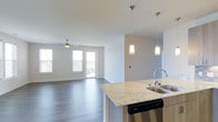 This single-floor residence at Overture offers a thoughtfully designed open-concept layout that seamlessly connects the kitchen and living areas. The kitchen is appointed with elegant granite countertops, a stainless steel sink and dishwasher, and a breakfast bar peninsula that doubles as a casual dining space. Stylish pendant lighting hangs above the counter, adding a modern touch to the space. Rich, dark hardwood-style flooring extends throughout the main living area, providing a warm contrast to the light, neutral-toned walls. The spacious living area is bathed in natural light, courtesy of large windows and glass doors that lead to a private balcony, offering views of the surrounding area. A ceiling fan ensures year-round comfort in the living space. The overall ambiance is clean, contemporary, and welcoming, with a bright and airy feel achieved through the combination of white ceilings, soft wall tones, and generous glazing. This well-appointed residence at Overture reflects a modern aesthetic with practical, high-quality finishes throughout.