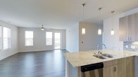 This single-floor residence at Overture offers a thoughtfully designed open-concept layout that seamlessly connects the kitchen and living areas. The kitchen is appointed with elegant granite countertops, a stainless steel sink and dishwasher, and a breakfast bar peninsula that doubles as a casual dining space. Stylish pendant lighting hangs above the counter, adding a modern touch to the space. Rich, dark hardwood-style flooring extends throughout the main living area, providing a warm contrast to the light, neutral-toned walls. The spacious living area is bathed in natural light, courtesy of large windows and glass doors that lead to a private balcony, offering views of the surrounding area. A ceiling fan ensures year-round comfort in the living space. The overall ambiance is clean, contemporary, and welcoming, with a bright and airy feel achieved through the combination of white ceilings, soft wall tones, and generous glazing. This well-appointed residence at Overture reflects a modern aesthetic with practical, high-quality finishes throughout.