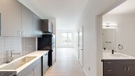 This thoughtfully designed 1-bedroom, 1-bathroom unit offers a clean, modern aesthetic throughout its efficiently arranged single-floor layout. Light-toned hardwood flooring flows seamlessly from room to room, complementing the crisp white walls and creating a bright, cohesive atmosphere.

The kitchen is outfitted with sleek gray cabinetry, white countertops, and a subway tile backsplash in a soft gray tone. Black stainless appliances — including a refrigerator, electric range, and over-the-range microwave — provide a bold contrast against the lighter finishes, while a single-basin sink with a modern faucet adds practical functionality.

The living area is bathed in natural light thanks to large sliding windows that frame outdoor views of surrounding greenery. The space feels open and airy, with recessed lighting and clean architectural lines reinforcing the contemporary design.

The generously sized bedroom features its own wall-mounted mini-split HVAC unit for personalized climate control, along with large windows that invite additional natural light and outdoor views.

The bathroom maintains the unit's modern character with a dark-toned vanity, clean white walls, matte black hardware accents, and warm wood-look flooring. A dedicated laundry area houses a stacked Energy Star-rated washer and dryer, adding everyday convenience. The unit is accessed through a solid, secure entry door, completing this well-appointed living space.