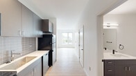 This thoughtfully designed 1-bedroom, 1-bathroom unit offers a clean, modern aesthetic throughout its efficiently arranged single-floor layout. Light-toned hardwood flooring flows seamlessly from room to room, complementing the crisp white walls and creating a bright, cohesive atmosphere.

The kitchen is outfitted with sleek gray cabinetry, white countertops, and a subway tile backsplash in a soft gray tone. Black stainless appliances — including a refrigerator, electric range, and over-the-range microwave — provide a bold contrast against the lighter finishes, while a single-basin sink with a modern faucet adds practical functionality.

The living area is bathed in natural light thanks to large sliding windows that frame outdoor views of surrounding greenery. The space feels open and airy, with recessed lighting and clean architectural lines reinforcing the contemporary design.

The generously sized bedroom features its own wall-mounted mini-split HVAC unit for personalized climate control, along with large windows that invite additional natural light and outdoor views.

The bathroom maintains the unit's modern character with a dark-toned vanity, clean white walls, matte black hardware accents, and warm wood-look flooring. A dedicated laundry area houses a stacked Energy Star-rated washer and dryer, adding everyday convenience. The unit is accessed through a solid, secure entry door, completing this well-appointed living space.