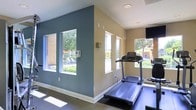 This single-story fitness space is designed to provide a functional and comfortable workout environment. The room features a combination of slate-blue and warm beige walls, creating a visually balanced and energizing atmosphere. Multiple windows along both walls flood the space with abundant natural light while offering views of an outdoor pool and landscaped surroundings, enhancing the overall workout experience.

The vaulted ceiling adds a sense of openness and airiness to the room, preventing it from feeling confined despite its focused purpose. Recessed lighting supplements the natural light, ensuring the space remains well-lit at all times. The flooring appears to be a smooth, easy-to-clean surface well-suited for exercise equipment.

Fitness equipment visible in the space includes a treadmill and what appears to be an additional cardio machine, positioned to take advantage of the outdoor views through the windows. A wall-mounted television is also present, providing entertainment during workouts. The clean white baseboards and well-maintained walls contribute to the tidy, purposeful ambiance of this fitness facility.