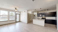This single-story, 1-bedroom, 1-bathroom unit offers a thoughtfully designed open-concept layout that maximizes the available living space. Light wood-style flooring runs consistently throughout, creating a clean, cohesive aesthetic. The living area is bright and airy, featuring a ceiling fan, a front entry door with transom window, and large windows that invite natural light and outdoor views. The kitchen is a standout feature, boasting rich dark cabinetry that extends to the ceiling for ample storage, light granite-style countertops, a central island with a sink, pendant lighting, and black appliances including a range, microwave, and refrigerator. A hallway with louvered double doors provides convenient closet storage and connects the main living areas to the private quarters. The bedroom is a comfortable retreat with a ceiling fan, two windows overlooking the exterior, and easy access to the adjacent full bathroom. The bathroom is well-equipped with a wide vanity featuring dark cabinetry and granite-style countertops, a large mirror, and a tiled tub-shower combination. The overall ambiance is clean, modern, and move-in ready.