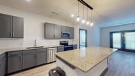 This single-floor unit showcases a cohesive modern design carried consistently throughout every room. The kitchen serves as a central highlight, featuring gray shaker-style cabinetry, granite countertops, and a full suite of stainless steel appliances including a refrigerator, electric range, microwave, and dishwasher. A generously sized kitchen island with matching granite provides additional prep space and casual seating. Four geometric pendant lights suspended from a track mount above the island add a contemporary industrial touch, complementing the light wood-look flooring that runs through the main living areas.

The bedrooms offer plush carpeting, neutral walls, and natural light from well-positioned windows with blinds. A ceiling fan in one bedroom adds both function and comfort. Each room features gray-trimmed doorways that tie into the overall color palette.

The bathrooms maintain the same design language, with gray cabinetry, granite vanity tops, and wood-look flooring. The primary bathroom includes a dual-sink vanity, a large mirror, a walk-in shower, and direct access to a walk-in closet. A secondary bathroom provides a single-sink vanity with similar finishes and a separate shower. The overall ambiance is clean, contemporary, and move-in ready.