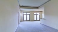 This single-floor space makes a strong first impression with its generous, open-plan layout and notably elevated ceilings adorned with elegant crown molding and a contrasting upper accent band, adding architectural depth and visual interest to the room. The living area is expansive and airy, finished with soft, neutral-toned wall-to-wall carpeting that adds warmth underfoot.

Two sets of French doors with transom windows above dominate the far wall, flooding the interior with abundant natural light while providing access to what appears to be an outdoor balcony or terrace. The large glass panels create a seamless visual connection to the exterior and contribute to the bright, open ambiance of the space.

To the right, a partial view of a kitchen or bar area is visible, featuring a countertop overhang that serves as a transitional element between the living area and the adjoining space. A pendant light fixture hints at a dedicated dining or kitchen zone nearby.

The overall aesthetic is clean and neutral, with warm beige walls, white trim, and subtle architectural detailing that lend the space a polished, welcoming character well-suited to a variety of interior styles.