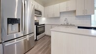 The York Suite is a thoughtfully designed 2-bedroom, 1-bathroom residence located in London, Ontario. Throughout the unit, light grey wood-style flooring and neutral warm-toned walls create a clean, contemporary aesthetic that flows seamlessly from room to room. The open kitchen is a standout feature, equipped with stainless steel appliances including a full-size refrigerator, range, and built-in microwave. Crisp white cabinetry, a classic white subway tile backsplash, and a light stone countertop with peninsula provide both style and functionality. The living room is bright and airy, with sliding glass doors opening onto a private balcony that offers sweeping views of the London city skyline. Both bedrooms are comfortably sized with large windows that bring in natural light and frame city views, complemented by the same consistent flooring and neutral palette. The primary bedroom offers a generous footprint well-suited for a full furniture arrangement. The bathroom features elegant marble-style surround tiling in the tub-shower combination, a modern vanity with stone countertop, and neutral floor tiles, delivering a polished, spa-inspired feel. The York Suite combines modern finishes with practical layout and urban views for a refined living experience.