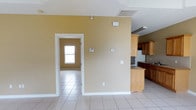 This single-story space features an open, functional layout finished with large, light-colored ceramic tile flooring that runs consistently throughout the main living areas. The walls are painted in a warm golden-beige tone, complemented by crisp white baseboards and trim that frame doorways and transitions between spaces. Ceiling-mounted HVAC vents indicate a central air system, contributing to practical comfort throughout.

The kitchen area is positioned to one side and includes a mix of medium-toned wood cabinetry alongside darker wood lower cabinets, providing ample storage. A countertop peninsula partially separates the kitchen from the adjacent living area, creating a natural boundary while maintaining an open feel. Upper cabinets extend toward the ceiling, maximizing vertical storage space.

A doorway leads to an additional room visible in the background, which features a window that allows natural light to filter into the space. The overall layout benefits from good connectivity between rooms, with wide doorways and an unobstructed flow. The neutral color palette and tile flooring give the space a clean, versatile foundation suited to a variety of uses.