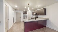 This single-floor residence showcases a thoughtfully designed open layout with a strong emphasis on modern finishes. The kitchen serves as the focal point, anchoring the living space with a striking deep burgundy island topped with speckled granite countertops and an integrated sink. Pendant lighting hangs above the island, adding warmth to the workspace. Dark espresso cabinetry lines the walls, complementing the full suite of stainless steel appliances, including a refrigerator, range, microwave, and dishwasher. Light wood-style flooring extends throughout the kitchen and into the living area, which benefits from natural light filtering through vertical blinds on a sliding door. The living space is open and versatile, offering a clean canvas for furnishing. The bedroom features plush carpeting and a window fitted with horizontal blinds, providing a quieter, softer retreat. The bathroom maintains the cohesive design language of the unit, with wood-style flooring, a dark vanity with granite countertop, and a full tub-shower combination with a curved curtain rod. Recessed lighting throughout the unit keeps the overall ambiance bright and contemporary.