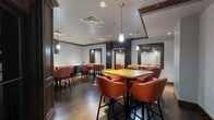 Located at 200 North Harvey Avenue in downtown Oklahoma City, this versatile lounge space offers a well-appointed, multi-functional layout across a single floor. The interior is thoughtfully divided into distinct zones, each serving a unique purpose while maintaining a cohesive aesthetic throughout.

The dining area features rich hardwood flooring, dark wood trim, and crown molding that lend a classic, polished character to the space. High-top tables paired with upholstered barrel-back stools in a bold orange-and-red patterned fabric create a lively yet comfortable atmosphere. Pendant lights hang above the tables, providing warm, focused illumination.

A built-in kitchenette anchors one end of the dining zone, equipped with dark wood cabinetry, a continuous countertop, a sink, and a microwave — ideal for refreshment service or light catering needs.

The media lounge area transitions to a more relaxed setting, featuring plush sectional sofas and individual chairs in a deep orange tone, accented by golden yellow ottomans. A large wall-mounted flat-screen television serves as the focal point, while a dark drop ceiling with recessed lighting adds a theater-like ambiance underfoot by patterned carpeting.

A semi-private nook off the main circulation path offers an intimate round table with seating for small group conversations. Interior glass-paneled doors with dark wood frames connect the zones, allowing natural sightlines while maintaining defined areas throughout the space.