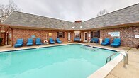 Located at 701 Green Mountain Dr in Little Rock, Arkansas, this two-floor property showcases a standout outdoor pool area that serves as a central amenity of the space. The generously sized rectangular swimming pool features striking blue tile accents along the waterline, stainless steel entry railings, and in-pool tanning ledges with reclined loungers — ideal for relaxation. The expansive concrete pool deck is lined with an abundance of blue lounge chairs, providing ample space for sunbathing and outdoor leisure. A separate hot tub or wading area with built-in reclined sun shelves adds an additional layer of comfort to the outdoor experience. The pool is enclosed by a wrought iron fence with decorative brick columns, offering both security and an open, airy feel that allows views of the surrounding landscape and tree line. The adjoining brick building features a hipped, shingled roof and dark shuttered windows, contributing a classic, residential architectural character to the setting. Outdoor lighting fixtures are mounted along the building's facade, extending the usability of the space into the evening hours. Tables and chairs are positioned within the fenced area, creating a comfortable outdoor gathering spot. The overall ambiance is relaxed and community-focused, well-suited for outdoor recreation and social enjoyment.