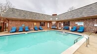 Located at 701 Green Mountain Dr in Little Rock, Arkansas, this two-floor property showcases a standout outdoor pool area that serves as a central amenity of the space. The generously sized rectangular swimming pool features striking blue tile accents along the waterline, stainless steel entry railings, and in-pool tanning ledges with reclined loungers — ideal for relaxation. The expansive concrete pool deck is lined with an abundance of blue lounge chairs, providing ample space for sunbathing and outdoor leisure. A separate hot tub or wading area with built-in reclined sun shelves adds an additional layer of comfort to the outdoor experience. The pool is enclosed by a wrought iron fence with decorative brick columns, offering both security and an open, airy feel that allows views of the surrounding landscape and tree line. The adjoining brick building features a hipped, shingled roof and dark shuttered windows, contributing a classic, residential architectural character to the setting. Outdoor lighting fixtures are mounted along the building's facade, extending the usability of the space into the evening hours. Tables and chairs are positioned within the fenced area, creating a comfortable outdoor gathering spot. The overall ambiance is relaxed and community-focused, well-suited for outdoor recreation and social enjoyment.