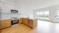 Located at 6320 Shakespeare Street in Pittsburgh's East Liberty neighborhood, this thoughtfully designed one-bedroom, one-bathroom unit offers a clean, modern aesthetic throughout. Light-toned hardwood flooring flows across the open living area, creating a warm and cohesive feel. The open-concept kitchen is a standout feature, anchored by a peninsula with warm wood cabinetry, gold-toned hardware, and a white quartz countertop. A subway tile backsplash, stainless steel appliances — including a range, microwave, and dishwasher — and recessed lighting complete the well-appointed cooking space. A large sliding window in the living area fills the room with natural light and frames views of the surrounding Pittsburgh skyline and greenery. The bedroom is a bright, serene retreat with light wood flooring, crisp white walls, and a window overlooking the building exterior. The full bathroom features large-format gray tile flooring, a walk-in glass-enclosed shower with white subway tile, and a sleek wood vanity with gold hardware and an illuminated mirror. A private balcony with composite decking and metal railing offers sweeping views of the city and the building's outdoor pool area below, making it an ideal spot for taking in the Pittsburgh landscape.