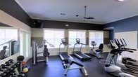 This single-floor fitness room offers a practical and comfortable environment for exercise. The space is outfitted with multiple treadmills positioned along a row of windows fitted with white blinds, allowing ample natural light to fill the room while providing a view of surrounding greenery. A recumbent bike and additional strength-training equipment round out the cardio and resistance offerings. The walls feature a two-tone color scheme with a dark horizontal band contrasting against lighter upper and lower sections, accented by white crown molding that adds a polished touch. Dark rubber flooring covers the ground, providing both durability and safety during workouts. A ceiling fan with an integrated light fixture hangs centrally, promoting air circulation throughout the space. Wall-mounted televisions are positioned for easy viewing during cardio sessions. The overall ambiance is clean, functional, and well-maintained, making it a well-rounded fitness facility suited for a variety of exercise routines.