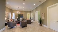 This single-story space features an open, generously proportioned layout that exudes a welcoming and polished ambiance. The main living area is defined by notably high ceilings adorned with crisp crown molding and bright white trim, lending an air of sophistication. Recessed lighting is distributed throughout the ceiling, complemented by an elegant chandelier that serves as a focal point and adds a refined touch to the space.

The flooring consists of large-format neutral tile, providing a clean and durable surface that ties the room together visually. Walls are finished in a warm olive-green tone, creating a grounded, earthy atmosphere. Seating is arranged thoughtfully, with upholstered chairs and sofas positioned around area rugs to define distinct conversation zones. Decorative potted plants are placed throughout, introducing natural elements and a sense of freshness.

Natural light filters in through windows dressed with patterned curtains, while wall-mounted artwork adds visual interest. Additional doorways visible along the back wall suggest connecting rooms, contributing to the functional flow of the overall layout.
