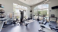 Located at 20 Pointe of Woods Dr N in Sayreville, New Jersey, this well-appointed fitness center offers a bright and energizing environment for a complete workout experience. The space is defined by its soaring ceilings and expansive floor-to-ceiling windows, including a striking arched window wall, which flood the interior with abundant natural light and provide open views of the surrounding outdoor area, including a neighboring pool and landscaped grounds.

The exercise room is equipped with a comprehensive selection of cardio machines, including treadmills, ellipticals, and stationary bikes from brands such as Star Trac. Strength training needs are met with a multi-station cable machine, free weights, dumbbells on a dedicated rack, resistance bands, and exercise balls. A wall of mirrors along one side enhances the sense of space while supporting proper form during workouts.

The flooring is a durable, speckled rubber gym surface designed for both comfort and safety. Ceiling fans are suspended throughout to maintain air circulation, and a wall-mounted flat-screen television adds a convenient entertainment option. The overall ambiance is clean, modern, and welcoming, making this fitness center a functional and pleasant space for daily exercise.