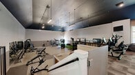 Located at 11001 W 133rd Ter in Overland Park, Kansas, this single-floor fitness center offers a generously sized, well-appointed exercise facility designed to support a comprehensive range of workout routines. The space is defined by its striking vaulted ceilings finished in a deep teal-black tone, which contrast boldly against bright white walls accented with a grey tile wainscoting. Sleek pendant and linear lighting fixtures illuminate the floor throughout, creating a focused yet energizing atmosphere. The main workout area is divided into functional zones, each with purpose-specific flooring. Durable rubber flooring anchors the strength training section, which is outfitted with a wide array of resistance machines, free weights, a dumbbell rack, benches, and a dedicated artificial turf lane for functional and sled-based training. A separate carpeted section houses cardio equipment, including treadmills, ellipticals, stair climbers, and rowing machines, with large windows that invite natural light and outdoor views of the surrounding greenery. Wall-mounted mirrors and a flat-screen television enhance the cardio area. A comfortable lounge area with upholstered armchairs and a round coffee table provides a welcoming space for rest and recovery. Stairs connect the main floor to an elevated mezzanine-style platform housing additional cardio machines.