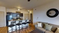 Located at 2200 Bellamy Cir in Greenville, North Carolina, this single-floor residence offers a well-designed, open-concept layout that seamlessly connects the kitchen and living areas. The kitchen is outfitted with dark cabinetry, black appliances including a microwave and range, and a generous countertop that extends into a breakfast bar with seating for multiple guests — an ideal setup for casual dining or entertaining. Track lighting above the kitchen provides focused illumination, while a ceiling fan in the living area helps maintain a comfortable atmosphere throughout. The warm-toned hardwood-style flooring flows continuously across the main living space, adding a cohesive and inviting aesthetic. The living area is comfortably sized, offering ample room for seating and relaxation. Neutral wall tones keep the interior feeling bright and versatile. A hallway visible from the main living space leads to additional areas of the home, suggesting a thoughtful and functional floor plan. The overall ambiance is cozy yet practical, making this space well-suited for everyday living.