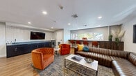 Located at 630 Pershing Blvd in Reading, PA, this single-floor communal lounge space offers a thoughtfully designed environment for gathering and relaxation. The interior is anchored by warm, light-toned hardwood flooring that flows seamlessly throughout the living room and hallway, complemented by recessed lighting that creates a bright and inviting atmosphere.

The living room serves as the heart of the space, featuring a large wall-mounted flat-screen television set against a striking geometric patterned tile accent wall. Beneath it, a run of sleek navy cabinetry with a wet bar and sink provides a functional and stylish entertaining station, complete with a mini-fridge. Vibrant burnt-orange tufted barrel chairs are paired with a rich brown leather sectional sofa accented with teal and yellow throw pillows, centered around a marble-top coffee table — creating a warm and eclectic seating arrangement.

A built-in counter-height bar with bar stools lines one wall beneath a row of windows, allowing natural light to filter in and offering views of the surrounding neighborhood. The walls are painted in soft gray-blue tones, adding depth to the space. Decorative artwork and accent furnishings round out the interior, giving the lounge a polished, contemporary feel well-suited for community use.