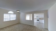 This single-floor residence offers a bright, open layout designed for comfortable everyday living. The main living area is finished with neutral-toned carpet and painted in soft, light tones that create an airy atmosphere. A distinctive arched window on one wall allows natural light to pour into the space, complementing the flush-mount ceiling light overhead. The living area flows seamlessly into the kitchen through an open pass-through cutout in the wall, maintaining a connected feel between the two spaces. The kitchen features white cabinetry with ample storage, light-colored countertops, and a white electric range. Tile flooring in the kitchen provides a practical contrast to the carpeted living area. Vertical blinds on the kitchen window offer light control while maintaining brightness. The overall palette of whites and neutrals throughout both areas contributes to a clean, cohesive aesthetic. The layout is straightforward and functional, making efficient use of the available space while ensuring the living and cooking areas remain visually connected without sacrificing definition between the two zones.