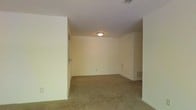 This single-floor space presents a straightforward, functional layout centered around a generously sized carpeted room visible in the image. The walls are finished in a light, neutral cream tone, contributing to a bright and open atmosphere despite the absence of windows in the immediate view. Overhead, a flush-mount ceiling light fixture provides consistent illumination throughout the room, while HVAC supply and return vents are positioned both on the ceiling and near the baseboard, indicating a full climate control system. The flooring throughout is wall-to-wall carpet in a warm beige tone, adding a layer of comfort underfoot. A partial wall or column is visible in the foreground, suggesting a defined entryway or transitional area leading into the main room. Electrical outlets are noted along the walls, and the overall condition of the space appears well-maintained and vacant, making it readily adaptable for a variety of uses. The clean lines, neutral palette, and practical infrastructure of this space provide a versatile foundation for customization.