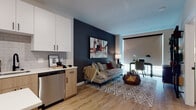 This thoughtfully designed single-floor residence combines modern aesthetics with functional living across its open layout. Light wood-tone flooring flows continuously throughout, creating a cohesive and warm foundation for the entire space.

The kitchen is a standout feature, showcasing two-tone cabinetry with crisp white upper cabinets and natural wood lower cabinets, paired with quartz countertops and a classic subway tile backsplash. A central island with bar seating adds workspace and casual dining convenience, while a full suite of stainless steel appliances — including a gas range, microwave, dishwasher, and refrigerator — ensures modern functionality.

The open-concept living and dining area is anchored by a bold dark accent wall that adds depth and drama to the space. Large windows with roller shades allow for abundant natural light, and the area comfortably accommodates a dedicated workspace alongside a well-furnished lounge area.

The bedroom features a rich burgundy accent wall that creates a warm, intimate atmosphere, complemented by a roller shade window for light control. The full bathroom is finished with a wood-toned vanity, quartz countertop, matte black fixtures, and a tiled soaking tub and shower combination, with striking horizontal stripe wall detailing adding a distinctive design touch.