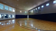 This indoor basketball court in Spring, TX is a well-appointed athletic facility designed for serious play and recreational use alike. The court features a polished hardwood floor with clearly marked regulation lines in red, blue, and black, including three-point arcs, lane markings, and center court designations. Soaring ceilings provide ample vertical clearance, while wall-mounted, glass-backboard basketball hoops are positioned at each end of the court. The walls are finished in a two-tone scheme — navy blue on the lower half and taupe on the upper — creating a clean, athletic aesthetic. A row of clerestory windows runs along the upper perimeter of the walls, allowing natural light to filter in while maintaining a focused interior environment. Recessed ceiling vents and overhead lighting ensure consistent illumination throughout the space. One side of the court opens visually to an adjacent room featuring large floor-to-ceiling windows and a second-floor overlook with a railing, offering a spectator viewing area. Multiple entry doors with exit hardware are accessible from the court floor. The overall atmosphere is functional, spacious, and purpose-built for athletic activity.