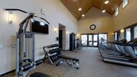 This fitness center is a thoughtfully designed workout facility that combines functionality with an inviting architectural aesthetic. The room's most striking feature is its soaring vaulted ceiling, clad in warm wood paneling and accented by recessed lighting, which lends an open and airy atmosphere to the space. Neutral-toned walls and carpeted flooring complement the warm tones overhead, creating a comfortable environment for exercise.

The facility is comprehensively equipped to support a variety of workout routines. A full dumbbell rack with a range of free weights is paired with an adjustable workout bench, catering to strength training needs. A wall-mounted flat-screen television provides entertainment during workouts. Multiple treadmills are positioned along one side of the room, while a cable resistance machine and additional strength equipment occupy the central area.

Large glass double doors on the far end of the room allow generous natural light to filter in, connecting the interior to the outside environment. A classic analog wall clock keeps members on schedule. Wall sconces provide supplemental ambient lighting throughout. The overall design balances practical fitness amenities with a polished, welcoming aesthetic suited to daily use.