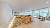 This elegantly designed amenity space spans a single floor and offers a refined, multipurpose environment suited for both relaxation and collaboration. The layout flows seamlessly from one zone to the next, unified by wide-plank light hardwood flooring, crisp white walls, and recessed lighting that maintains a bright, airy atmosphere throughout.

The main lounge area is anchored by a generously sized, curved sectional sofa in warm peach bouclé fabric, set atop a richly patterned area rug. A wall-mounted display, subtle LED cove lighting, and built-in shelving with greenery add warmth and character. A secondary seating nook features cane-back accent chairs and bench seating beneath floating shelves lined with potted plants, creating an inviting retreat.

A long, white marble-topped bar counter with wood-and-metal stools serves as a central gathering point, illuminated by a pair of striking pendant light fixtures. A curated gallery wall adds artistic interest to the space.

Floor-to-ceiling arched grid windows flood the interior with natural light and frame open exterior views, while a glass-enclosed meeting room with a round table and pendant light offers a semi-private setting for focused gatherings. Bar-height seating along the window-facing perimeter provides additional informal areas to work or socialize. Together, these elements create a polished, community-oriented space with a warm, contemporary aesthetic.