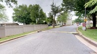 This single-story property is located along a calm residential street characterized by mature deciduous and evergreen trees that provide ample shade and a sense of established neighborhood greenery. A prominent privacy fence with lattice detailing runs along the property boundary, offering seclusion from the street. The surrounding area features well-maintained landscaping, with lush grass lining the sidewalk and shrubs visible near neighboring structures. The paved road leading past the property is wide and accessible, with clearly marked yellow lines indicating traffic flow. In the background, multi-story neighboring buildings are visible through the tree canopy, suggesting a moderately dense residential setting. The overall ambiance of the exterior is quiet and private, with the combination of mature trees and fencing contributing to a sheltered feel. The natural surroundings and greenery give the property a calm, suburban character well-suited to comfortable single-floor living.