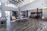 This single-story clubhouse in Clarksville, Indiana, is designed as a dynamic community gathering space that blends modern architectural design with versatile, amenity-rich interiors. The main lounge area is immediately striking, featuring dramatic vaulted ceilings with exposed dark wood beam accents, globe pendant lighting, and a bold teal accent wall that adds a contemporary energy to the open layout. Durable wood-look flooring runs throughout, tying each area together cohesively.

The social and recreational zone is well-equipped, offering ping pong tables, a shuffleboard table, and a large flat-screen TV alongside comfortable lounge seating — making it an ideal space for both casual gatherings and organized events. Floor-to-ceiling glass walls and double doors open directly to an outdoor pool area, flooding the interior with abundant natural light and seamlessly connecting indoor and outdoor recreation.

The reception area near the entrance features a welcoming front desk, built-in shelving, and direct sightlines to the exterior through large glass entry doors. A fully equipped kitchen serves the space with rich dark wood cabinetry, granite-style countertops, a white subway tile backsplash, stainless steel appliances, a generous island with bar seating, and an adjacent casual dining area with natural light from nearby windows. Together, these spaces create a well-appointed, community-focused clubhouse suited for a wide range of social activities.