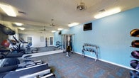 This fitness center offers a comprehensive workout environment designed to support a wide range of exercise routines. The space is finished with light blue accent walls that create an energizing yet calm atmosphere, complemented by a textured ceiling and durable patterned carpet flooring in grey and blue tones that adds both visual interest and practicality underfoot.

The layout is open and well-organized, providing ample room to move freely between equipment stations. A full-length wall mirror runs along one side of the room, visually expanding the space and allowing users to monitor their form during workouts. Ceiling fans ensure consistent air circulation throughout.

The equipment selection is well-rounded, featuring rowing machines positioned along the floor for cardio training, a multi-station cable weight machine for versatile strength exercises, and a dedicated free weights area stocked with a variety of kettlebells neatly arranged on a rack. A wall-mounted flat-screen television is positioned for easy viewing during workouts.

Natural light filters into the space through a doorway, while overhead fluorescent lighting ensures the entire area remains bright at all times. The overall design balances functionality with a clean, modern aesthetic, making this fitness center a practical and inviting space for daily exercise.