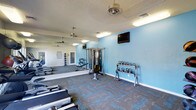 This fitness center offers a comprehensive workout environment designed to support a wide range of exercise routines. The space is finished with light blue accent walls that create an energizing yet calm atmosphere, complemented by a textured ceiling and durable patterned carpet flooring in grey and blue tones that adds both visual interest and practicality underfoot.

The layout is open and well-organized, providing ample room to move freely between equipment stations. A full-length wall mirror runs along one side of the room, visually expanding the space and allowing users to monitor their form during workouts. Ceiling fans ensure consistent air circulation throughout.

The equipment selection is well-rounded, featuring rowing machines positioned along the floor for cardio training, a multi-station cable weight machine for versatile strength exercises, and a dedicated free weights area stocked with a variety of kettlebells neatly arranged on a rack. A wall-mounted flat-screen television is positioned for easy viewing during workouts.

Natural light filters into the space through a doorway, while overhead fluorescent lighting ensures the entire area remains bright at all times. The overall design balances functionality with a clean, modern aesthetic, making this fitness center a practical and inviting space for daily exercise.