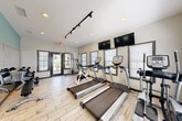 Located in Lithonia, GA, this single-room fitness center offers a well-equipped exercise space designed to support a well-rounded workout routine. The room features light wood-style flooring throughout, crisp white walls accented by a teal-painted corner, and recessed lighting complemented by a track lighting system, ensuring the space is bright and functional at any time of day. Multiple windows dressed with horizontal blinds line the walls, inviting natural light and providing views of the surrounding outdoor greenery. A glass-panel entry door reinforces the open, airy feel of the room. The cardio section is anchored by a row of TRUE-brand treadmills and an elliptical trainer, alongside a stationary spin bike, offering users a variety of aerobic options. Wall-mounted flat-screen televisions positioned above the cardio equipment allow for entertainment during workouts. A multi-function cable machine provides strength-training versatility, while a dedicated free-weights area includes a dumbbell rack with an adjustable bench. Full-length mirrors along one wall enhance the sense of space and allow users to monitor their form. Posted fitness center rules and a keypad entry system near the interior door reflect a managed and community-oriented environment. Overall, the space is thoughtfully arranged to maximize utility within a compact footprint.