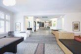 This single-floor lobby presents a thoughtfully designed communal space that seamlessly blends classic architectural details with contemporary furnishings. The entry vestibule welcomes visitors through glass doors, featuring large street-facing windows, light tile flooring, and a comfortable lounge chair paired with a statement floor lamp, setting an inviting tone.

The space is divided into several distinct zones connected by wide-open sightlines and architectural columns. Elevated terrazzo flooring defines a more intimate gathering area, accessed via broad granite steps, where a round dining table surrounded by modern chairs sits beneath globe pendant lights. Framed artwork lines the crisp white walls throughout, adding color and personality.

A lower-level lounge area features large format tile flooring, built-in bench seating, and an open layout that accommodates casual seating and a game table. A boldly painted teal-green accent wall anchors one section, providing striking contrast to the otherwise neutral palette.

Additional amenities visible include a reception desk area, mailboxes, fitness equipment, and a billiards table tucked within an alcove. Abundant natural light pours in through multiple windows, enhancing the airy, gallery-like ambiance throughout the entire space.