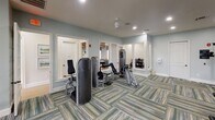 Located at 9325 Blue House Rd in North Charleston, SC, this fitness center is a thoughtfully designed, single-floor facility offering a comprehensive workout environment. The space is finished with soft blue-gray walls and distinctive multi-toned striped carpet tiles that lend a modern, polished aesthetic throughout.

The cardio zone is positioned along a wall of large picture windows, flooding the area with natural light and providing pleasant outdoor views of a neighboring deck and greenery. This section is equipped with treadmills, elliptical cross-trainers, and a recumbent bike, all featuring individual digital displays. Ceiling fans are installed overhead to maintain comfortable airflow during workouts.

The strength-training area is generously appointed with multiple cable and resistance machines, accommodating a wide range of muscle groups and fitness levels. Full-length mirrors line one wall, enhancing the sense of spaciousness and allowing users to monitor their form. Recessed lighting throughout ensures the entire space is well-illuminated.

French-style glass doors connect the fitness center to an adjacent community area, creating a seamless flow between amenity spaces. The overall ambiance is clean, bright, and welcoming, reflecting a commitment to providing residents with a high-quality, functional fitness experience.