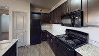 This single-floor residence offers a thoughtfully designed living space with a strong emphasis on modern style and functional layout. The kitchen stands out as a central feature, showcasing rich dark espresso cabinetry that extends from floor to ceiling, providing generous storage throughout. Light-toned granite-style countertops create a striking contrast against the dark cabinetry, lending the kitchen a polished and contemporary aesthetic. Black appliances, including a full-size refrigerator, electric range with coil burners, and an over-the-range microwave, complete the cohesive, moody color palette. The kitchen is finished with durable wood-look plank flooring that adds warmth and is easy to maintain. A built-in wine rack above the refrigerator adds a refined decorative touch. Neutral, light-toned walls keep the space feeling open and bright, while recessed ventilation ensures comfort. A paneled door visible from the kitchen hints at additional rooms within the residence. Overall, the space balances practicality with a modern design sensibility, making it both visually appealing and highly functional for everyday living.
