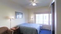 This single-floor space offers a clean, functional layout designed for comfort and ease. The bedroom serves as the centerpiece of the room, furnished with a generously sized bed dressed in a soft blue quilted spread, accompanied by a wooden nightstand for convenient bedside storage. Overhead, a white ceiling fan provides both ambient lighting and airflow, contributing to a comfortable atmosphere throughout the space. A wall-mounted sconce near the bed offers focused task lighting, adding to the room's practical design. The large window along one wall allows an abundance of natural light to flood the interior, creating a bright and airy feel. Blue carpeting anchors the room with a cool, calm tone that complements the neutral, light-colored walls. A framed artwork piece adds a decorative touch, enhancing the overall aesthetic without overwhelming the understated design. A compact heating and cooling unit is positioned beneath the window, ensuring year-round climate control. The overall ambiance is calm and straightforward, with thoughtful details that make the space both welcoming and highly functional for everyday use.