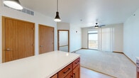 This single-floor residence in Rapid City, South Dakota, offers a thoughtfully designed open-concept layout that seamlessly connects the kitchen and living areas. The kitchen is a standout feature, showcasing warm medium-toned wood cabinetry that extends to the upper walls, a crisp white subway tile backsplash, a large center island with a clean white countertop and built-in storage, and a full suite of stainless steel appliances including a refrigerator, range, microwave, and dishwasher. Stylish pendant lights hang above the island, complementing the recessed ceiling lighting. Light wood-look flooring runs throughout the kitchen and entry areas, transitioning to plush carpet in the living and bedroom spaces. The living area is bright and open, anchored by a ceiling fan and a sliding glass door that leads to a private balcony with outdoor views. Multiple bedrooms offer comfortable proportions, carpeted floors, and ample natural light from well-placed windows. One bedroom includes access to a walk-in closet and an adjoining bathroom, which features a full tub-shower combination. The overall ambiance is clean, modern, and inviting, with warm wood tones and neutral walls creating a cohesive and comfortable living environment.