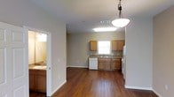 This two-floor residence features a practical and well-organized layout designed for comfortable everyday living. The main living area is anchored by rich, dark hardwood flooring that extends throughout, adding warmth to the neutral-toned walls and white trim detailing.

The kitchen is positioned at the far end of the main living space and is equipped with medium-toned wood cabinetry both above and below the counter, offering ample storage. A white dishwasher and refrigerator provide standard appliances, while a single-basin sink sits beneath a window that allows natural light to filter into the space. The countertops feature a stone-like finish that complements the cabinetry. Overhead, a flush skylight-style fixture and a hanging pendant light with a bowl-shaped shade provide layered lighting to the area.

Visible through a doorway to the left is an adjoining bathroom, which includes a vanity with a sink and warm overhead lighting, suggesting a functional and well-lit space. The clean white walls and cabinetry in this area contrast with the darker flooring found in the main living zone.

Overall, the space maintains a cohesive, understated aesthetic with consistent finishes and a functional floor plan across both levels.