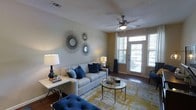 This single-floor space showcases a thoughtfully designed living room that blends comfort and contemporary style. The room is anchored by a plush neutral-toned sofa accented with deep navy blue throw pillows, complemented by a tufted blue accent chair that adds a bold pop of color. A round glass-and-gold coffee table serves as a refined centerpiece, positioned atop a gold and ivory abstract area rug that adds visual warmth to the dark hardwood flooring beneath.

The walls are painted in soft warm gray tones, providing a neutral backdrop that highlights the decorative sunburst mirrors mounted as a focal art arrangement. Ambient lighting is provided by elegant table lamps on either side of the seating area, while a ceiling fan with an integrated light fixture offers both function and style overhead.

Natural light flows generously through large windows fitted with horizontal blinds and accented by dark curtain panels, with a glass-paneled entry door further brightening the space. The overall ambiance is welcoming and polished, combining transitional design elements with practical everyday comfort in a cohesive and inviting arrangement.