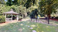 Located at 800 State St in West Columbia, SC, this well-maintained outdoor space offers a serene, park-like setting defined by mature trees, manicured lawns, and thoughtfully designed landscaping. A standout feature of the grounds is a classic white octagonal gazebo, framed by dense shrubs and towering trees that provide generous shade throughout the day. The gazebo serves as a natural focal point within the landscape and offers a sheltered spot for relaxation or gathering.

The grounds include a dedicated fenced area, which functions as a dog park, enclosed with black chain-link fencing and equipped with a gated entry and posted signage. This space provides a secure and designated area for pets to roam freely. Nearby, a concrete walking path winds through the property, lined with crape myrtle trees and neatly trimmed ornamental shrubs, creating an inviting corridor for strolling.

Mulched planting beds filled with low-growing shrubs and ground cover are interspersed throughout the grounds, adding texture and visual interest to the landscape. The overall ambiance is tranquil and green, with an abundance of natural canopy providing dappled light across the open lawn areas. The combination of open grass, structured pathways, and lush plantings creates a cohesive and well-maintained outdoor environment suited for leisure and community use.