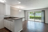 Located at 3700 S Plaza Dr in Santa Ana, CA, this efficiently designed single-floor unit makes excellent use of its space with a thoughtful, open layout. The combined kitchen and living room serves as the heart of the home, featuring rich wood-style flooring, a textured ceiling, and a kitchen peninsula topped with dark granite countertops. White cabinetry, stainless steel appliances—including a gas range and refrigerator—and a double stainless sink complete the functional kitchen. A pendant light fixture adds a decorative touch above the peninsula. The living area flows naturally toward a sliding glass door that fills the room with natural light and provides direct access to a private outdoor patio, where lush greenery creates a pleasant outdoor retreat. A separate bedroom area offers the same warm wood-style flooring and access to natural light through a vertical-blind-covered window. The full bathroom is neatly appointed with a vanity featuring white cabinetry, a mirror with overhead lighting, and a bathtub with a glass enclosure. Two closets provide ample storage, and a laundry area is conveniently integrated into the bathroom. Neutral wall tones throughout create a clean, bright atmosphere well-suited to a variety of interior styles.