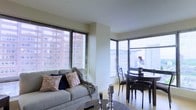 This single-floor urban residence offers a thoughtfully designed open-concept layout that seamlessly integrates the living and dining areas into one cohesive space. Large windows fitted with sleek horizontal blinds line multiple walls, flooding the interior with abundant natural light and framing views of the surrounding cityscape, giving the space a lively, connected-to-the-city ambiance.

The living area is anchored by a comfortable light grey sofa dressed with decorative patterned cushions and a cozy throw blanket, paired with a round coffee table styled with elegant pillar candle holders. The warm, light-toned flooring enhances the bright and airy atmosphere throughout.

Adjacent to the living area, the dining space features a dark-toned round table accompanied by matching dark wood chairs, offering an intimate setting for meals. A tasteful floral arrangement on the dining table adds a natural, welcoming touch.

The clean, neutral wall palette throughout the space provides a versatile backdrop that complements both the furnishings and the natural light pouring in from the corner window configuration. Overall, this space presents a modern, urban aesthetic with a warm and inviting character, well-suited for comfortable city living.