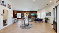 Located at 900 SE Third Ave in Ocala, FL, this single-floor space offers a well-appointed open-plan layout that seamlessly integrates a fully equipped kitchen with a generous living room area. Light wood-tone flooring runs throughout, lending warmth and visual continuity to the entire space.

The kitchen is outfitted with crisp white shaker-style cabinetry, granite countertops, stainless steel appliances including a refrigerator and dishwasher, and a peninsula island with bar stool seating beneath pendant lighting — ideal for casual gatherings. Local artwork adorns the walls, adding a sense of community identity.

The living area is expansive and thoughtfully zoned, featuring multiple seating arrangements that accommodate both intimate conversation and larger group settings. A bold, deep burgundy accent wall serves as a dramatic focal point, contrasting beautifully with the bright white walls and trim throughout. A wall-mounted flat-screen TV and an ornate mirrored console cabinet anchor this zone. Large windows dressed with white blinds fill the space with natural light and provide views of the surrounding greenery. Recessed lighting throughout ensures the space is well-lit at any hour. The overall ambiance is modern, welcoming, and polished — well-suited for a community lounge or clubhouse setting.